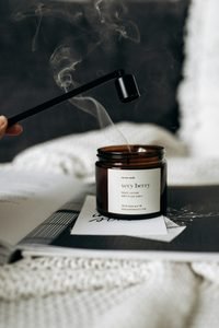 Brown glass candle jar labeled "very berry" on a magazine, with a black wick tool hovering above and wisps of smoke drifting up.